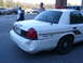 2003 Ford Crown Victoria - A white 2003 Ford Crown Victoria police car with a dented rear bumper is parked in a lot with other vehicles and people.