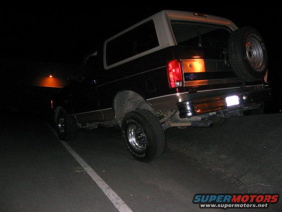 Rear view of a dark-colored Ford Bronco with a white top and a spare tire mounted on the back. It's parked at night