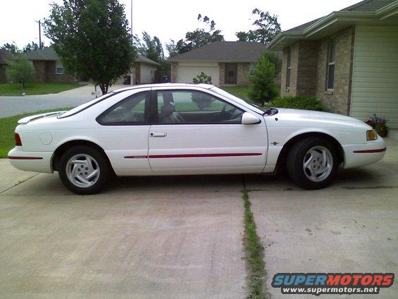 White coupe parked on a concrete driveway in front of a house. Red pinstripe along the lower body