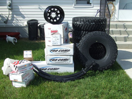 Stack of Pro Comp tires, a black wheel, and boxes of suspension parts on grass
