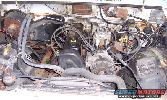 Engine bay with visible engine block, alternator, carburetor, and hoses. Some rust is present