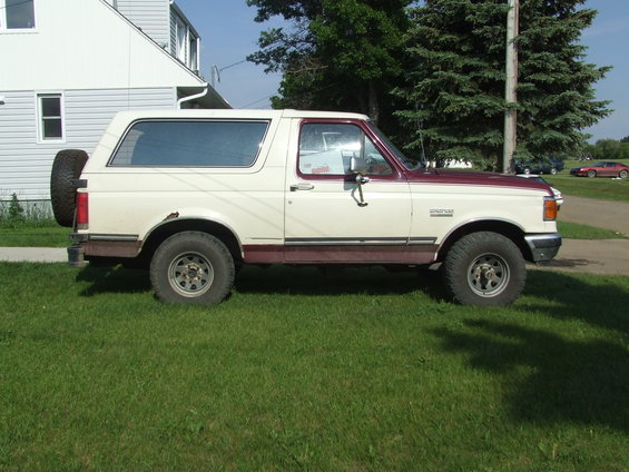 1990 Ford Bronco parked on grass, side view. Two-tone paint, spare tire on rear. House and trees in background