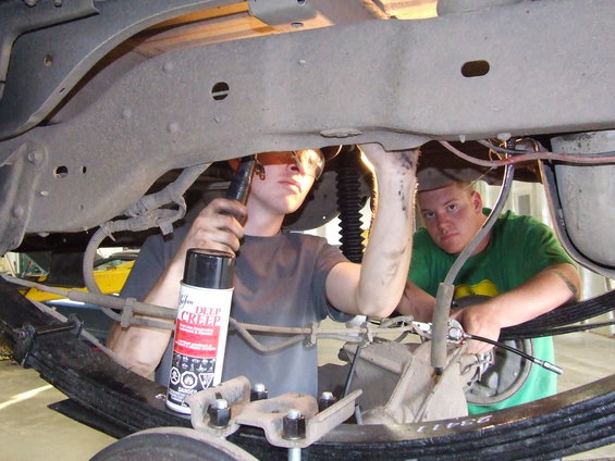 Undercarriage of a vehicle with two men working on it. One man sprays lubricant on suspension components