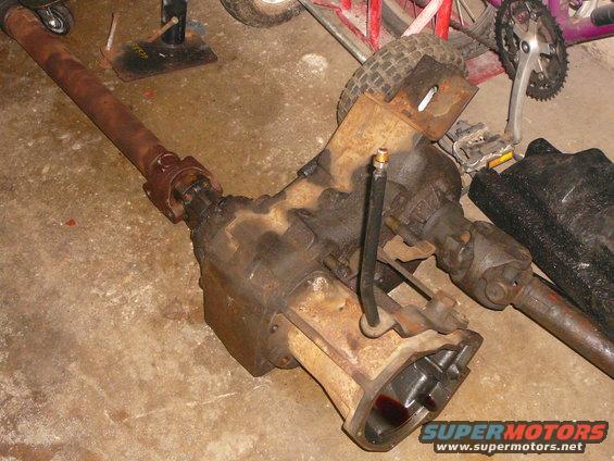 Transfer case and driveshaft assembly on a concrete floor. Bicycle parts and a tire are visible in the background