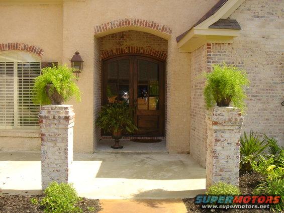 Front entrance of a house with double wooden doors, brick accents, and potted plants on pillars