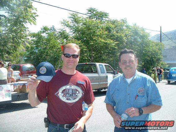 Two men stand in front of trees and vehicles at a parade. One man holds a baseball cap