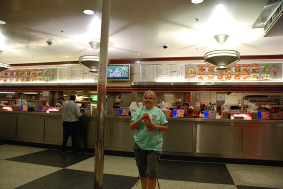 Interior of a fast food restaurant with menu boards and employees behind a counter. A woman stands in the foreground holding a red item