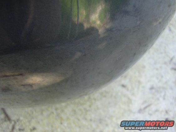 Close-up of a dark vehicle fender with reflections of trees and sky. Supermotors.net logo visible