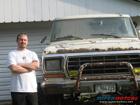 Guy in white t-shirt arms crossed next to a beat-up white Ford pickup. Truck has rust on hood and grille, yellow fog lights, and a brush guard