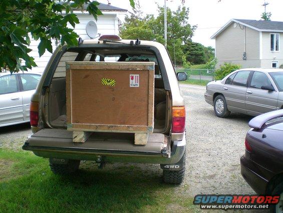 SUV's rear cargo area loaded with a large wooden crate; another sedan and a dark car are parked nearby