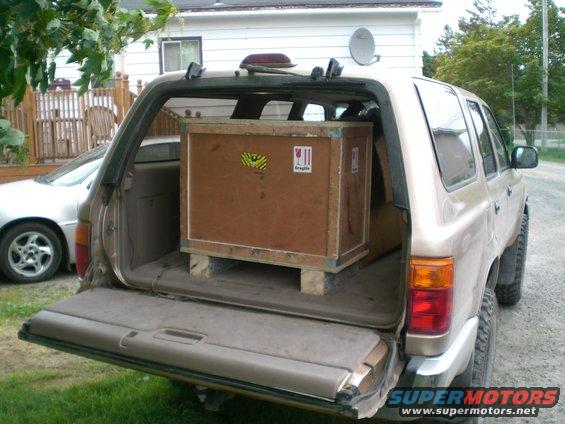 Tan SUV rear view with open tailgate and wooden crate inside. Another car is visible to the left