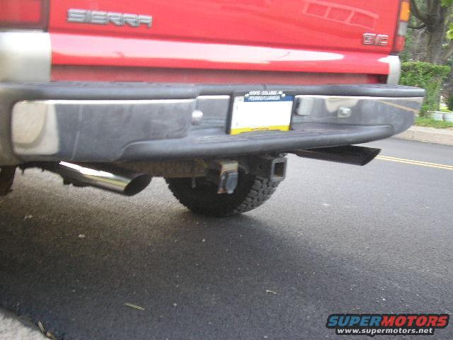Red GMC Sierra pickup truck rear bumper with dual exhaust pipes and a trailer hitch. License plate visible
