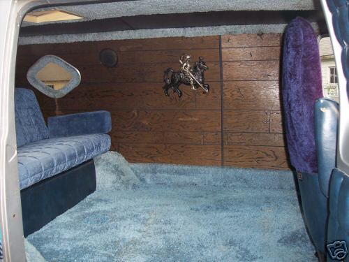 Interior of a van showing blue velour seats, blue carpet, wood-paneled walls, and a diamond-shaped mirror