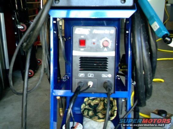 Blue welding machine with red power button and control knob, black cables attached