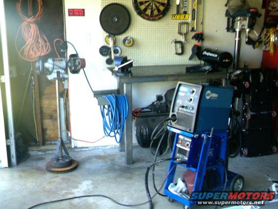 Welding machine on a blue cart, air compressor, and various tools on a pegboard wall in a garage