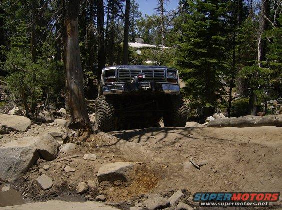Front view of a lifted, modified Ford Bronco on a rocky, wooded trail. Chrome grille, winch visible