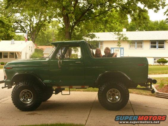 Green 1978 Ford Bronco, side view, with two passengers in the open top