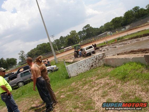 Mud bog event with people standing near cars and a tractor pulling a truck