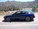 1995 Ford Probe - A blue 1995 Ford Probe GT is parked on a dirt road overlooking a lake and hills.