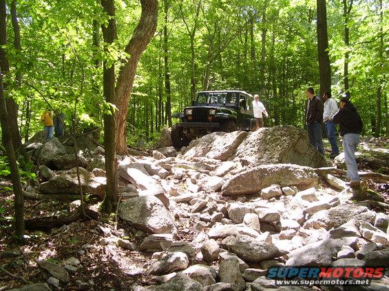 Black Jeep Wrangler navigating a boulder-strewn forest path, with several people observing