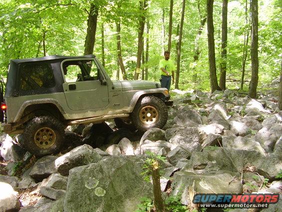 Jeep Wrangler driving over rocks in a wooded area. A person stands in the background