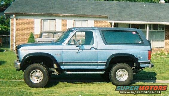 Light blue Ford Bronco parked in front of a brick house. Another vehicle is visible behind it