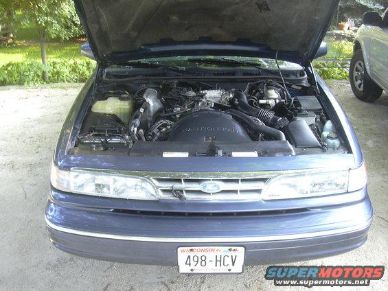 Blue Ford Crown Victoria with hood open showing engine. Another SUV is parked to the right