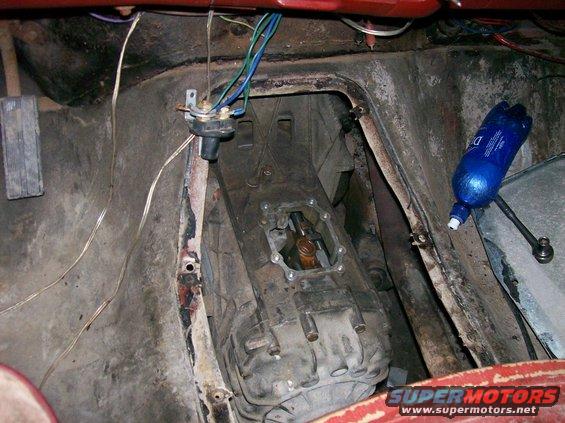 Interior shot of a vehicle's transmission tunnel, showing the gearbox and wiring. A blue bottle is visible on the right