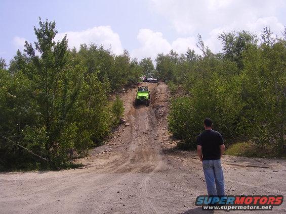 guy watching green off-road vehicle go up dirt hill with another vehicle behind it