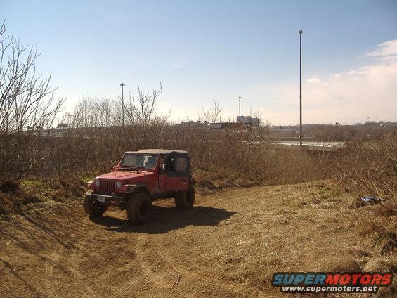 Red Jeep Wrangler on a dirt path with dry brush and a highway in the background
