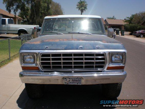 Front view of a blue 1978 Ford F-250 pickup truck with a rusty hood and grille. A white pickup truck is parked in the background