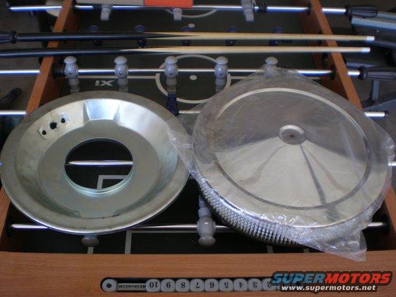 Air cleaner assembly parts on a foosball table. One part is a metal dish with a large central hole and three smaller holes. The other is a round filter in plastic wrap