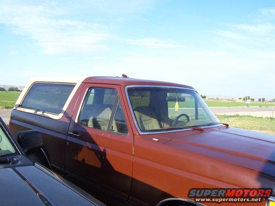 Side view of a brown Ford Bronco with a white top. Another dark vehicle is in the foreground