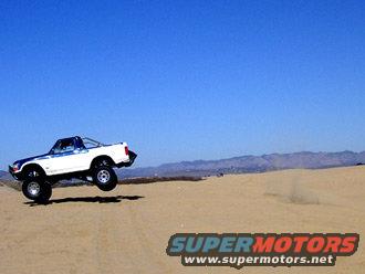 White off-road truck with blue stripe mid-air over sand dunes