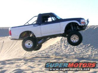 White and blue lifted pickup truck airborne over sand dune