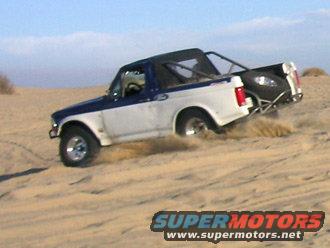 1996 Ford Bronco with blue top and white body driving on sand dunes, spare tire mounted on rear roll cage