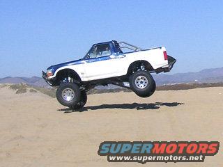 White and blue Ford Bronco airborne over sand dunes