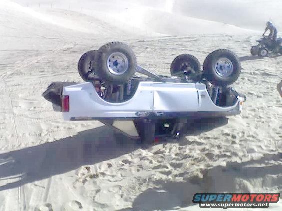White SUV overturned on sand dunes, showing its underside and wheels. A person rides an ATV in the background