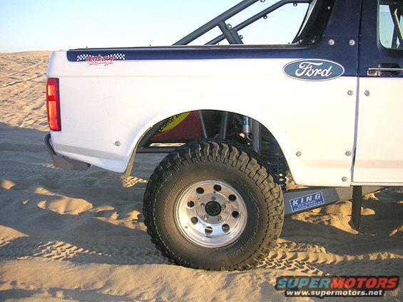 Side view of a white Ford Bronco prerunner with a blue stripe and roll cage, parked on sand
