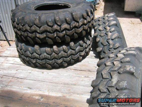Stack of four off-road tires on a wooden surface. One tire is on top of another, and two are to the right