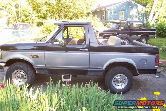 Side view of a two-tone black and silver Ford Bronco with a sun visor and aftermarket wheels. Another truck is parked behind it