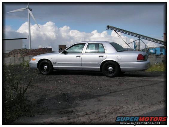 1998 Ford Crown Victoria parked on gravel with a wind turbine and industrial equipment in the background