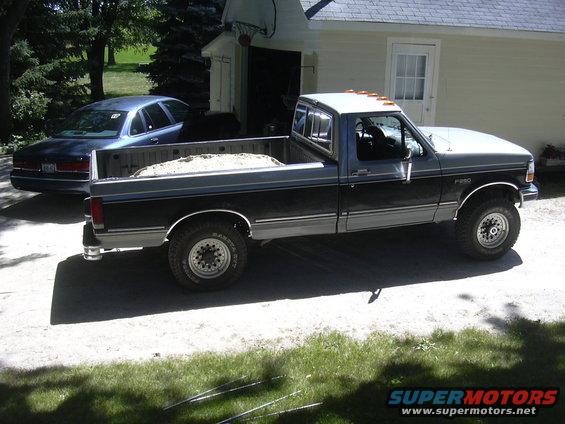 Gray and black Ford F-250 pickup truck parked in driveway with sand in bed. Blue sedan behind it