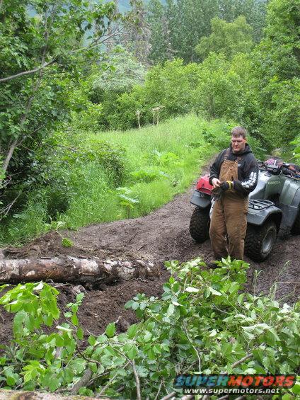 Man in overalls stands by a green ATV on a muddy path with a fallen log