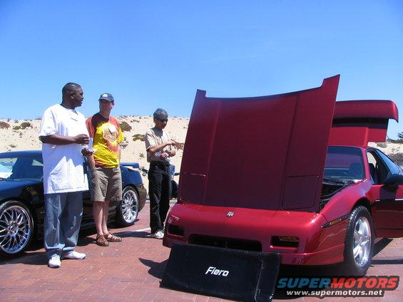 Red 1986 Pontiac Fiero with hood and doors open, parked next to a dark car. Three guys stand around