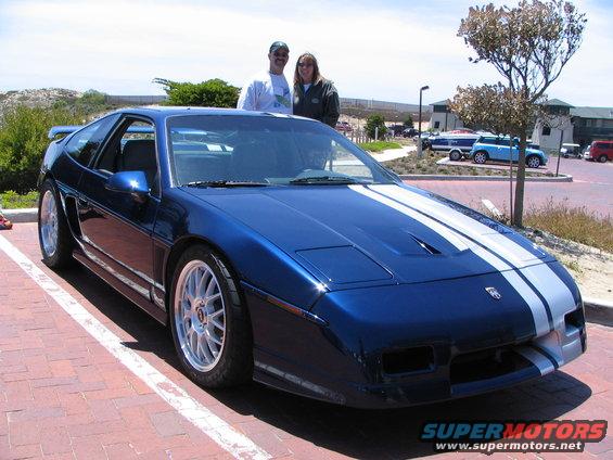 Blue 1986 Pontiac Fiero with white racing stripes parked on brick pavement, two people standing behind it