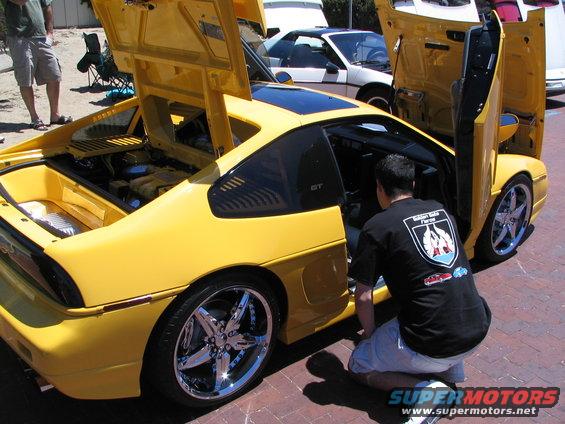 Yellow Pontiac Fiero GT with doors and hoods open, chrome wheels, man kneeling by driver's side
