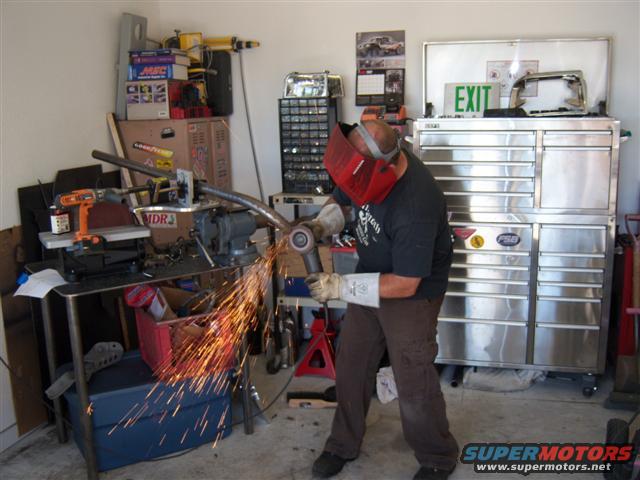 Man in red welding mask grinds metal tube with sparks flying, in a cluttered garage with tool chests and a whiteboard