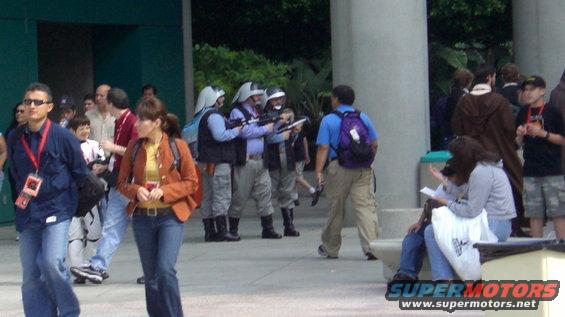People in costumes holding prop guns, some wearing helmets, in an outdoor area with other people around