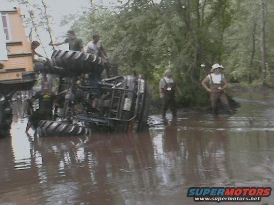 Overturned ATV in muddy water with people around, part of a yellow vehicle visible